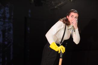 Zu sehen: Die Schauspielerin Marie Philipp in der Rolle des Kaufmannes Herr Michael Fischer. Sie trägt einen weißen Rollkragenpullover, eine schwarze Hose mit Hosenträgern und einen gelben Handschuh. In der rechten Hand hält sie ein Stock, in der linken ein Taschentuch. Sie guckt ängstlich zurück, während sie das Taschentuchan ihre Nase hält.