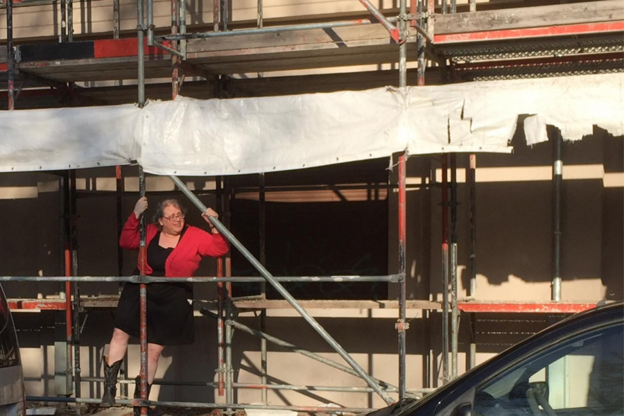 A white woman wearing a short black dress, coral cardigan sweater, black cowboy boots, and glasses is climbing up some scaffolding in front of a building. She is looking out to her left and smiling slightly, or maybe she is squinting in the afternoon soon. The edge of a white tattered tarp flaps behind her.