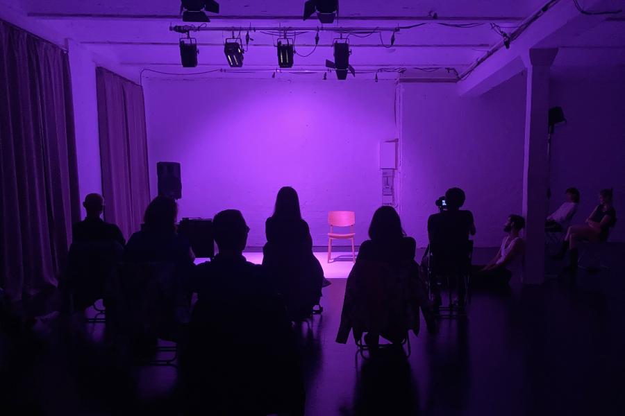 Aufnahme einer in Violett beleuchteten Bühne. Die Bühne, die hinter dem Publikum zu sehen ist, ist leer.