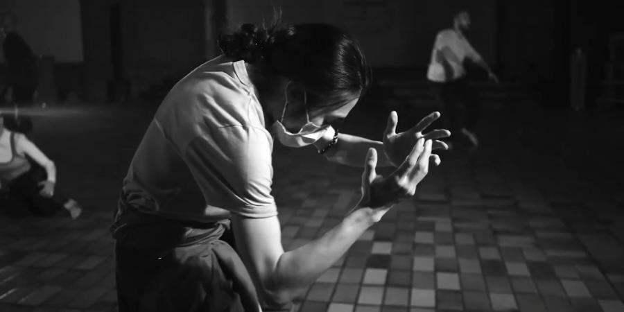 Gekrümmter Körper seitlich, der seine Hände in Richtung Gesicht hebt, das mit einer Maske bedeckt ist. Im Hintergrund unscharf ein tanzender Körper in der Kirche