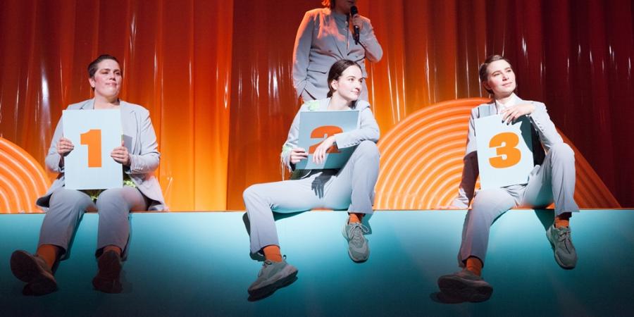 Drei Performerinnen in grauen Anzügen sitzen auf einem Bühnenrand und halten drei Tafeln mit den Nummer 1, 2 und 3 in den Händen. Im Hintergrund steht eine Frau vor einem orangefarbenen Vorhang und spricht in ein Mikro. 