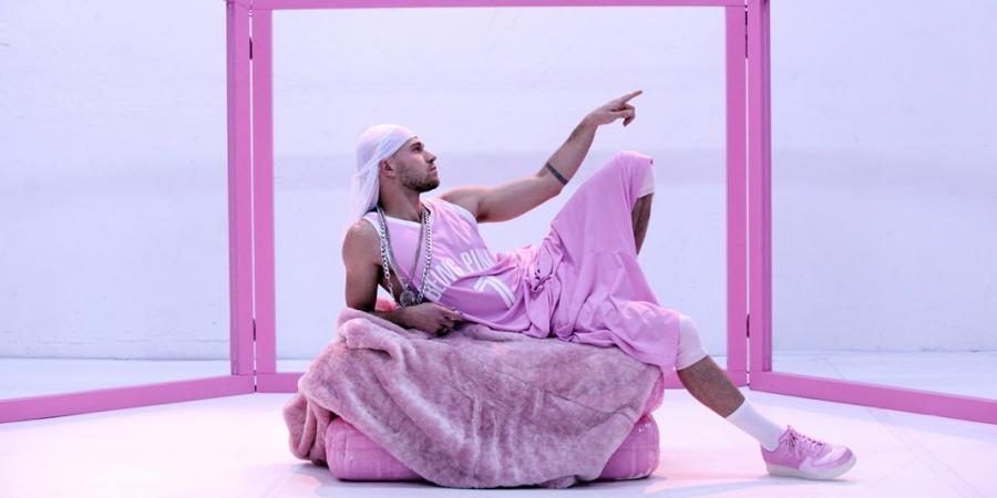 Ein Performer, der bekleidet ist mit einem weiten ärmellosen Trikot, einer rosafarbenen Sporthose und einem weißen Bandana, liegt auf einem rosafarbenen Polster. Sein linker Zeigefinger zeigt in die Ferne, sein linkes Bein ist angewinkelt. Die Pose ist Michelangelos Deckenfresko "Die Erschaffung Adams" nachempfunden. Im Hintergrund wird das Bühnenbild umrahmt von einem Gerüst aus rosafarbenen Streben. 
